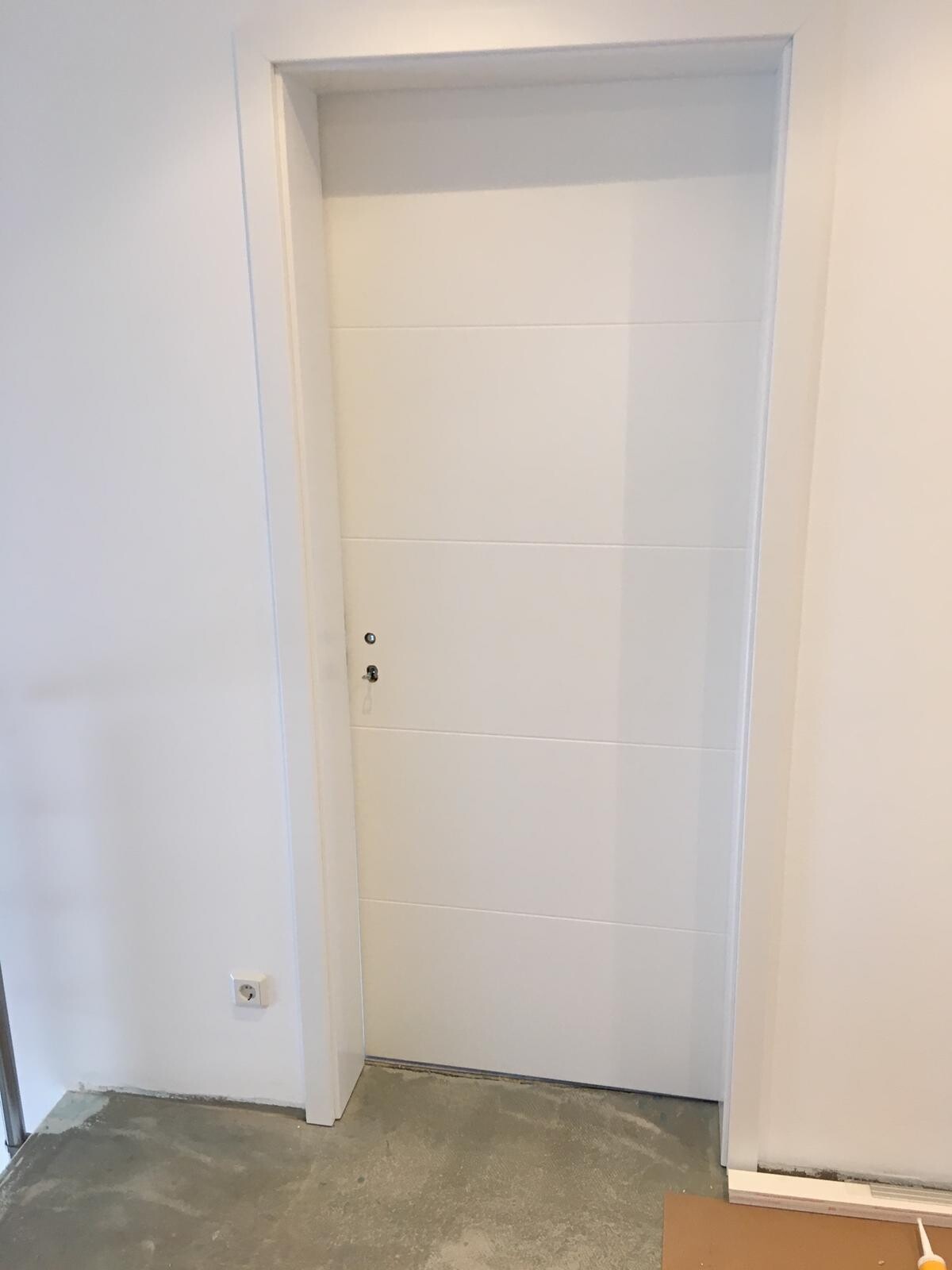 Weiße Tür mit vier Paneele in Innenraum; Türknauf sichtbar, Steckdose links, Boden unfertig.