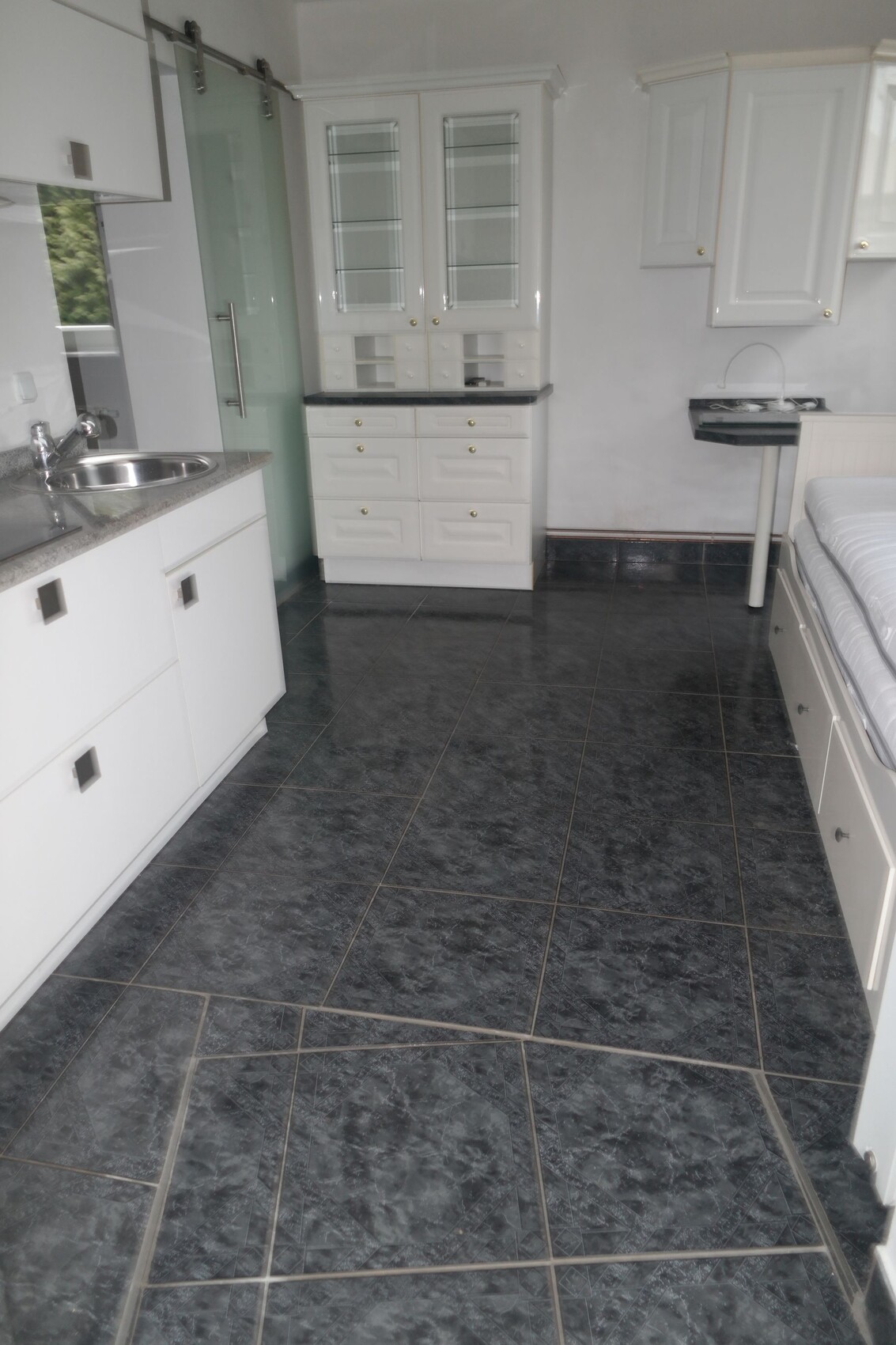 Weiße Küche mit Spüle, Glastür-Dusche und Bett rechts; dunkle Bodenfliesen.