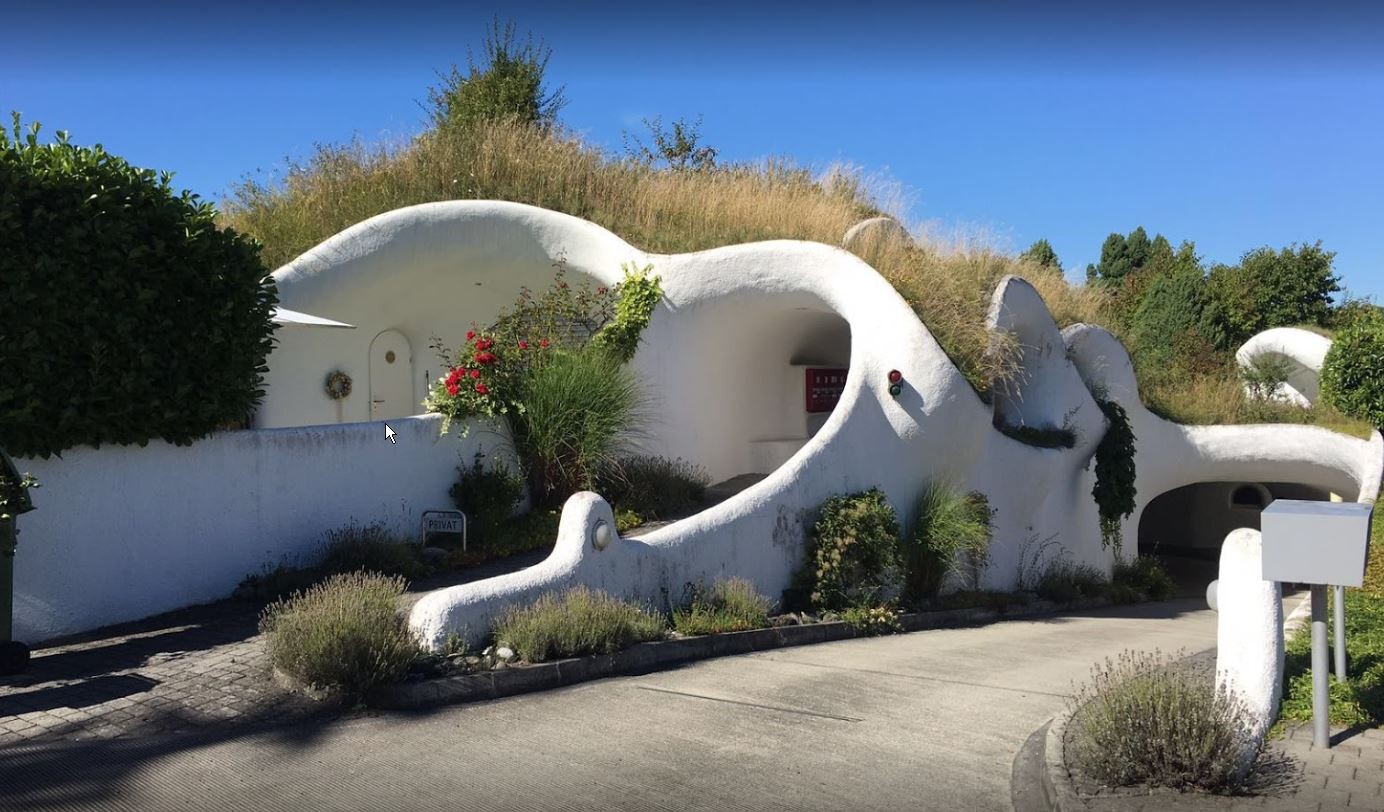 Weiße, kurvenreiche Wohnarchitektur mit bepflanztem Dach und Durchgangstunnel.
