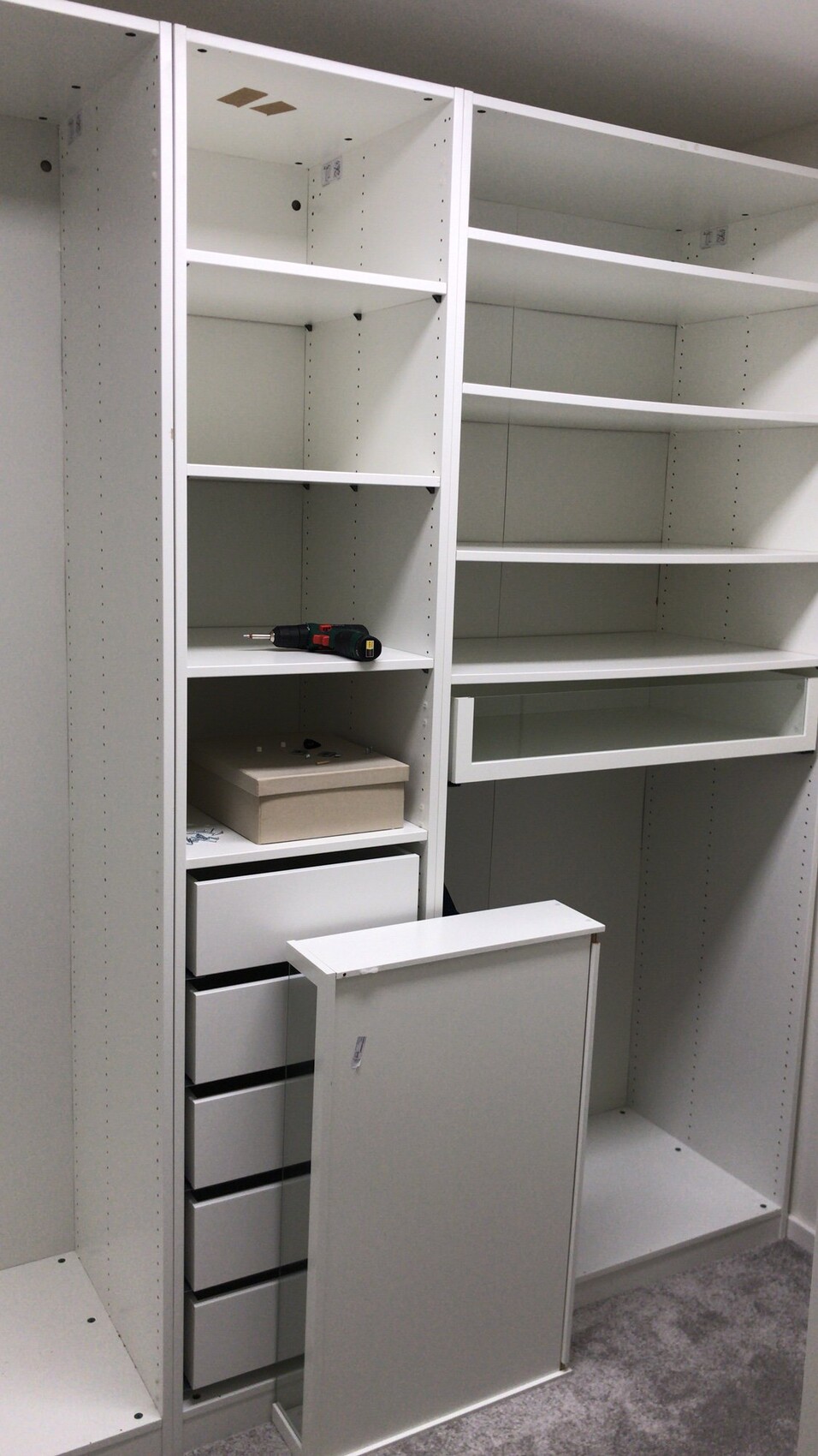 Weiße modulare Regale mit Schubladen; Bohrmaschine auf der Ablage, beige Box sichtbar.