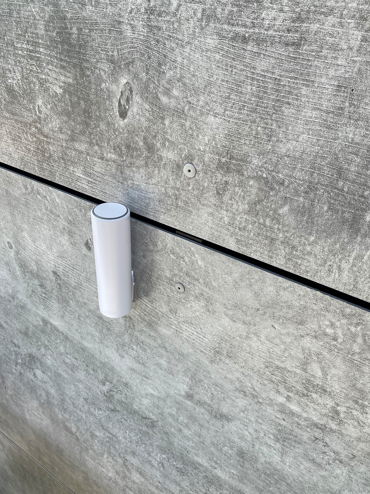 Weiße zylindrische Außenleuchte an grauer Betonwand, nahe einer horizontalen Fuge.