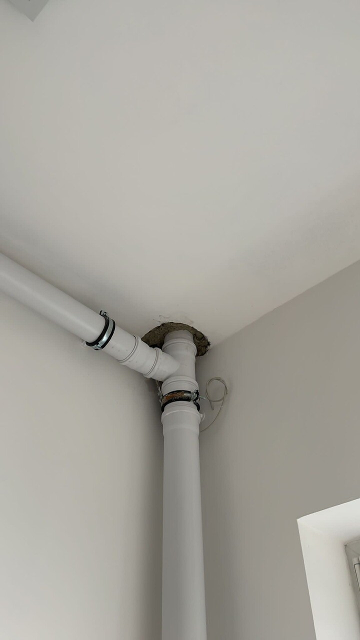 Weiße Kunststoffrohre mit Verbindungsstücken in einer Raumecke; Lochdurchbruch in Wand/Decke mit grobem Mörtel umrandet.