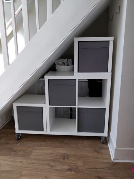Weiße modulare Aufbewahrungseinheit mit grauen Stoffboxen unter der Treppe auf Rollen.