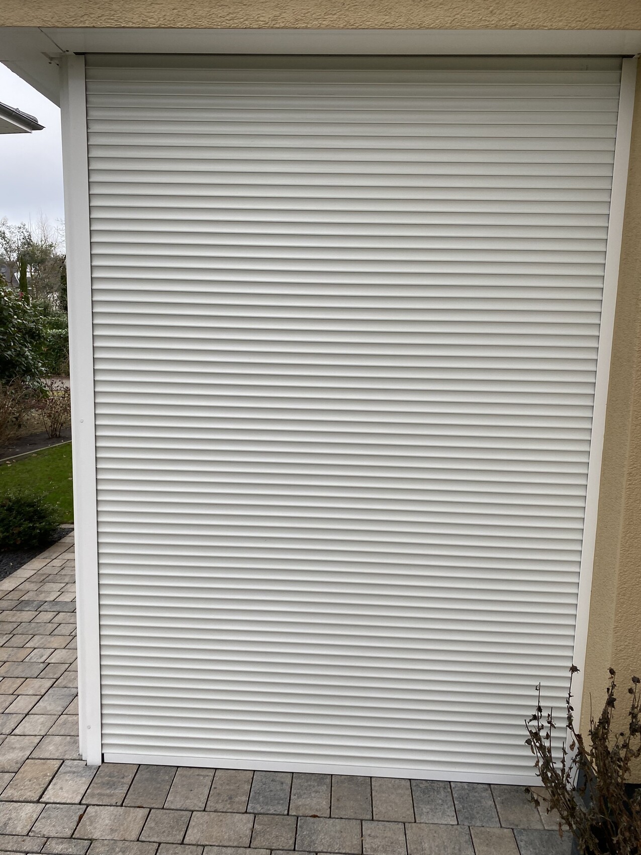 Weiße Rollladenfront vor beigeem Haus, gepflasterter Boden; Rollladen geschlossen.