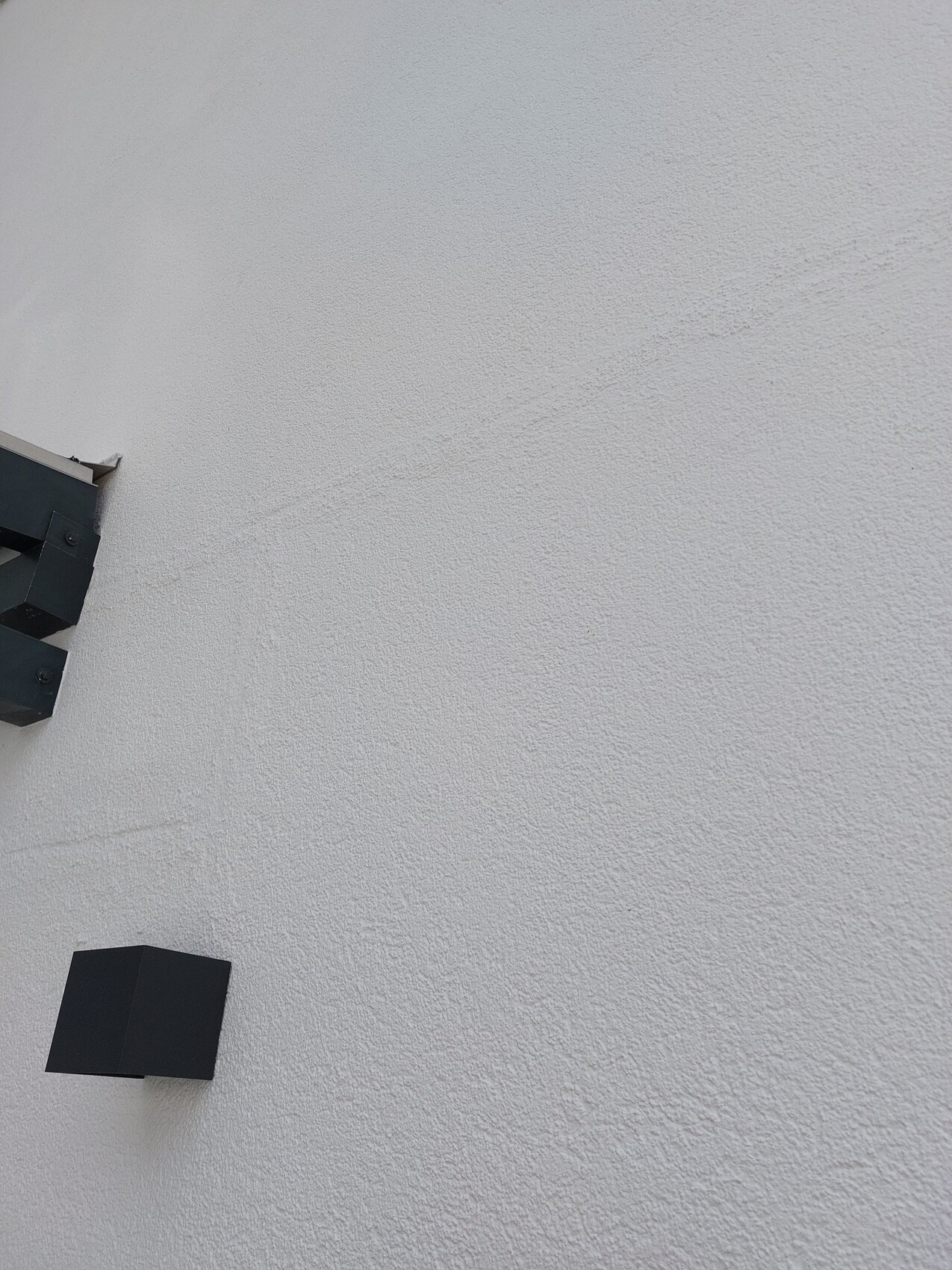 Weiße grob strukturierte Außenwand; links unten ein schwarzes, quaderförmiges Bauteil.
