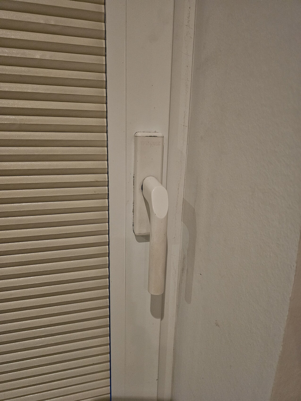 Nahaufnahme eines weißen Türgriffs an einer Tür, links eine beige Jalousie.
