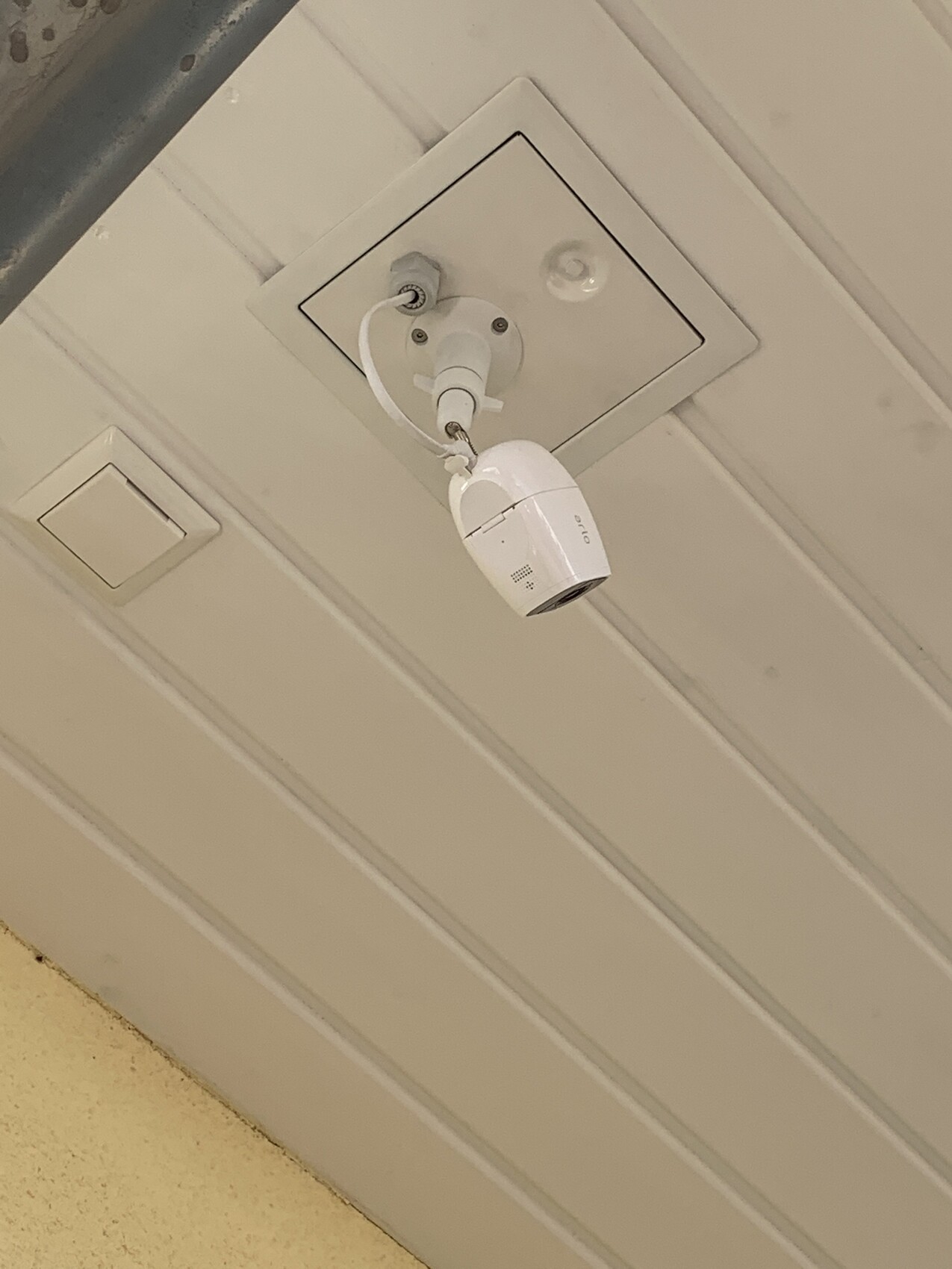 Kleine weiße Überwachungskamera hängt von der Decke neben einer quadratischen Abdeckung.