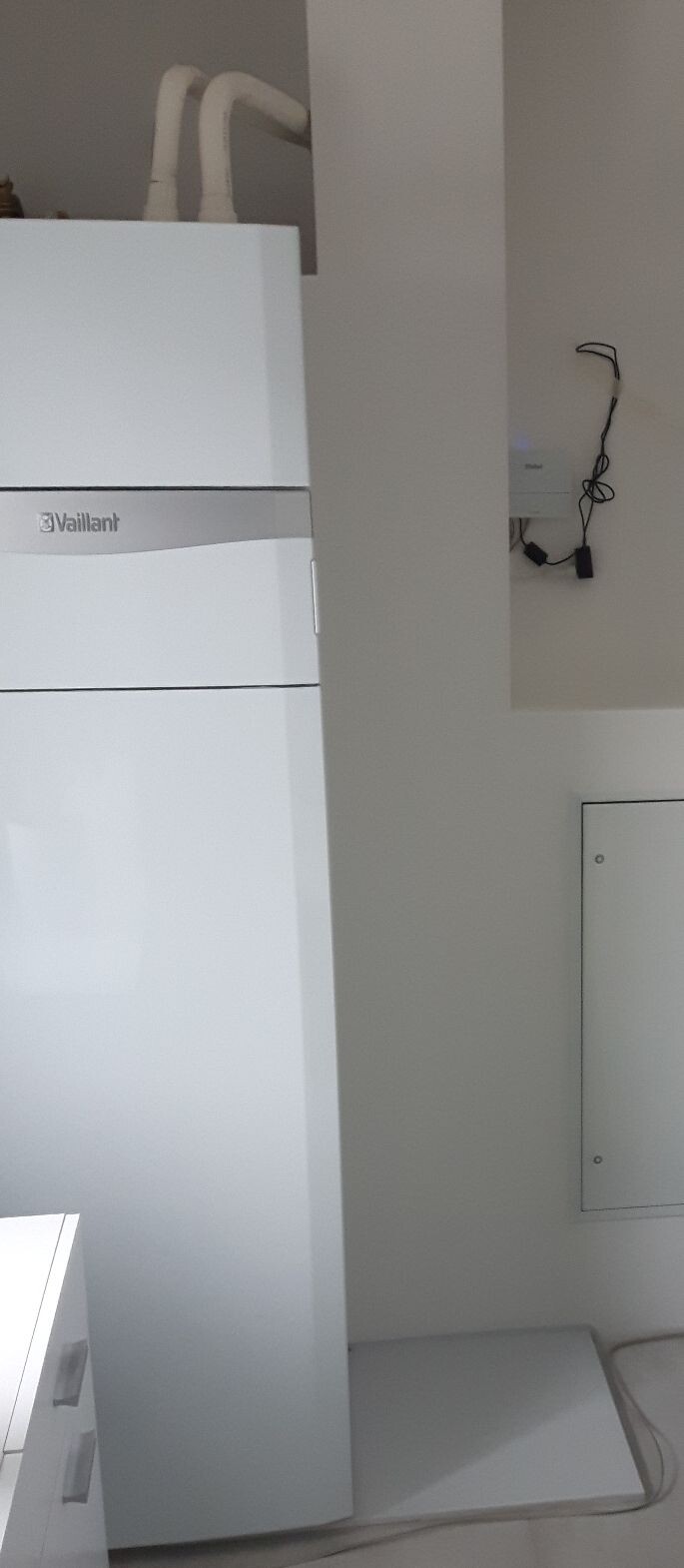 Weißer Vaillant Heizkessel in einem Kellerraum mit Rohren oben und Kabeln an der Wand.