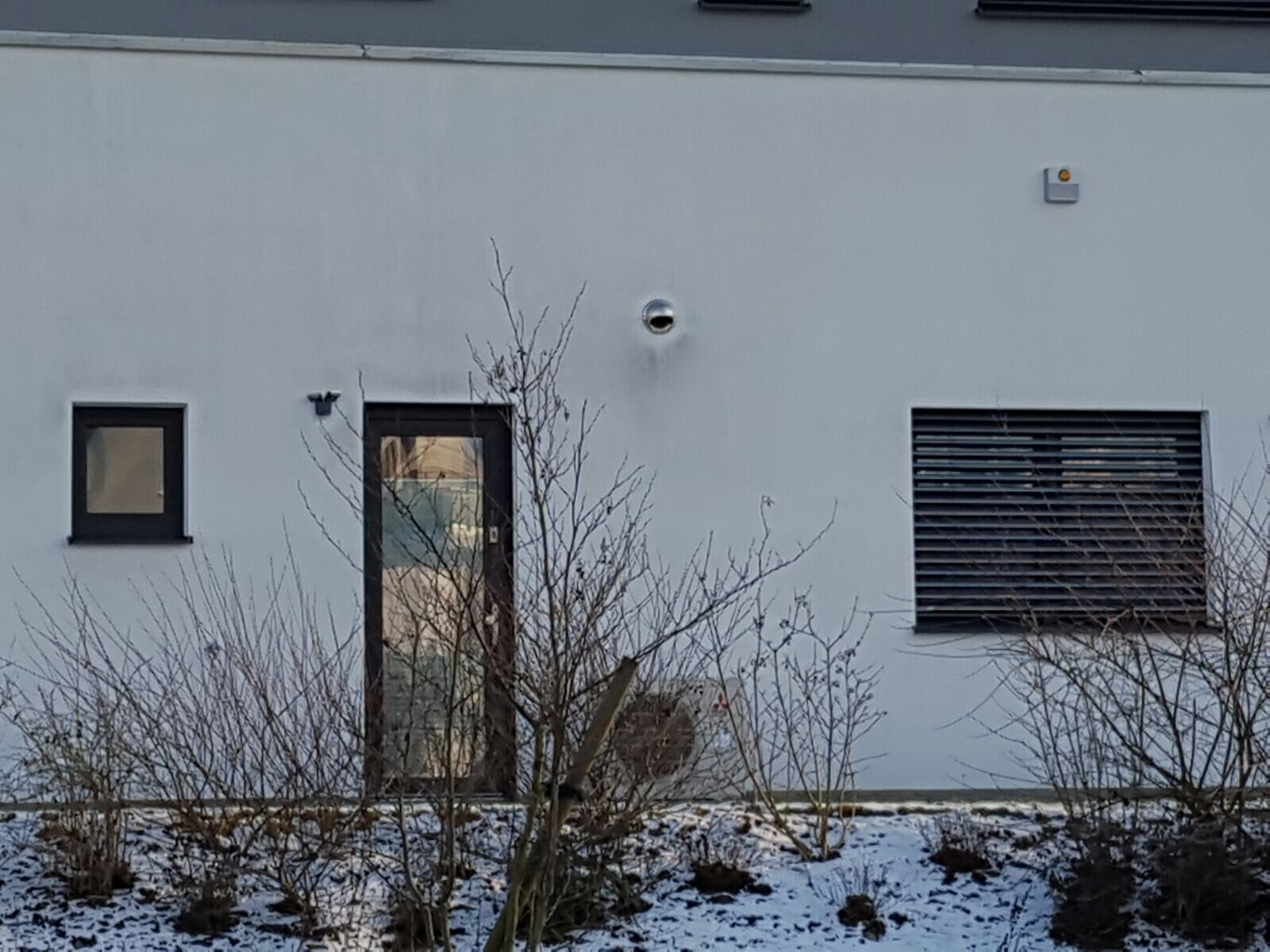 Außenwand eines Gebäudes: Tür mit Glas, Fenster, runde Lüftungsöffnung, Schnee und Sträucher.
