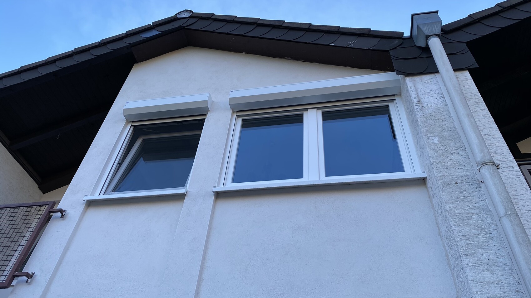 Weiße Hausfassade mit zwei Fenstern, dunklem Dach, Regenrinne und Fallrohr.