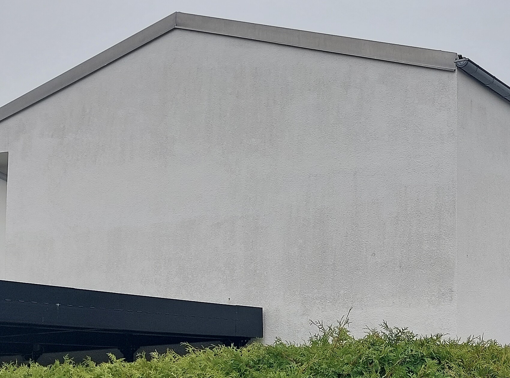 Graue verputzte Gebäudefassade mit geneigtem Dach, dunklem Vordach unten und grünen Büschen.
