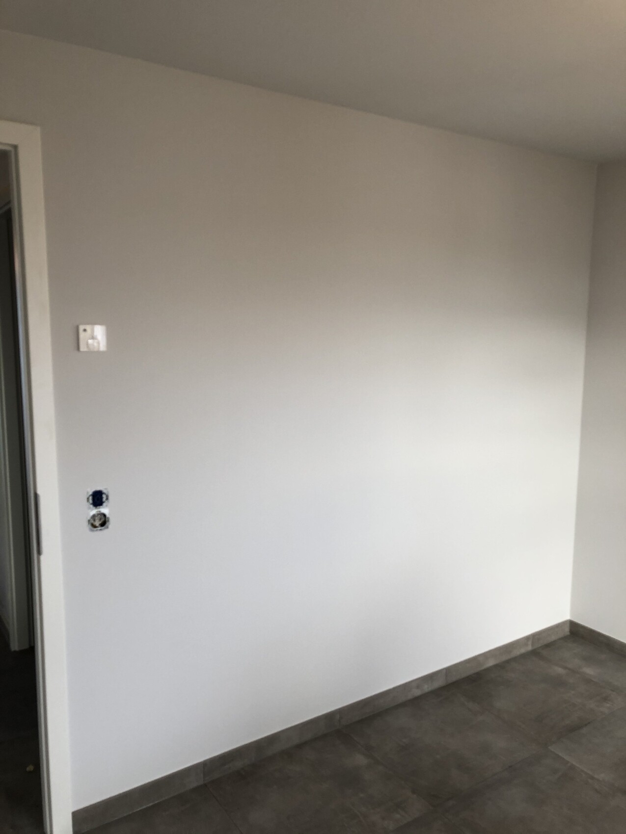 Leerer, heller Raum mit grauer Wand, Fliesenboden, Türrahmen links und Steckdosen.