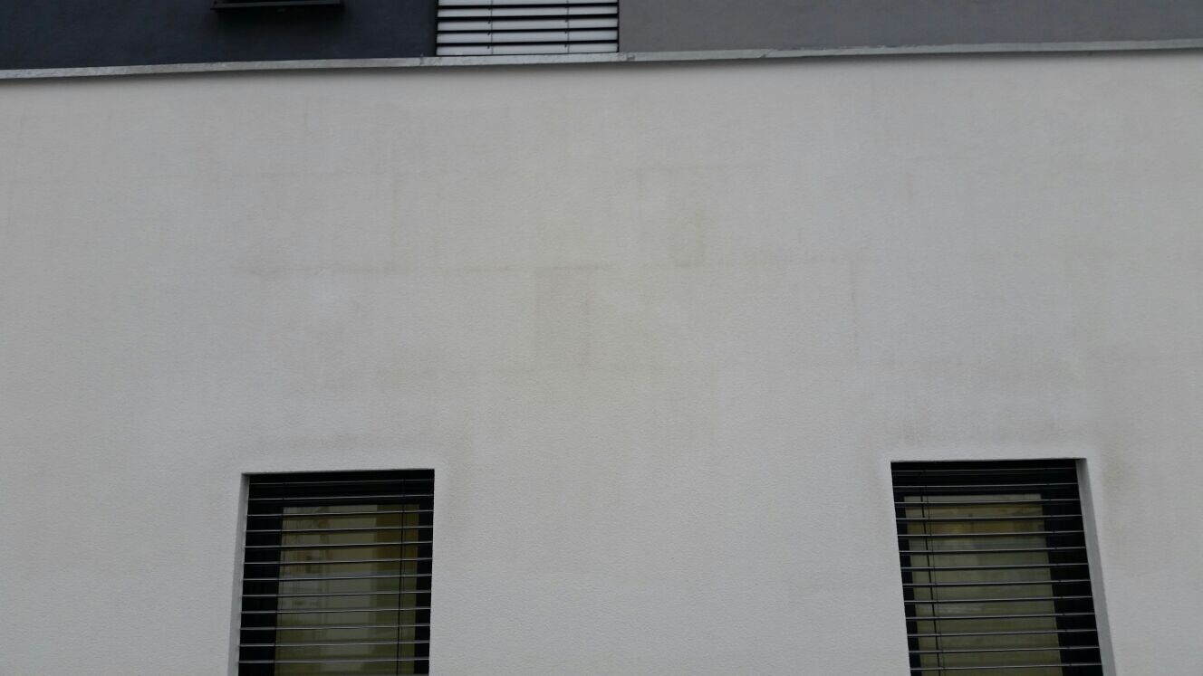 Weiße Außenwand mit zwei Fenstern und schwarzen Fensterläden, obere Belüftungsluke sichtbar.