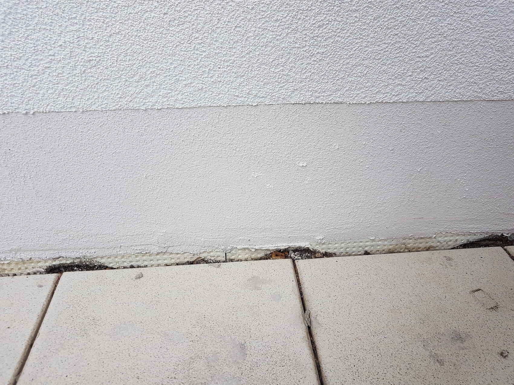 Weiße grobtexturierte Außenwand über hellen Fliesenboden; Sockel bröckelt leicht