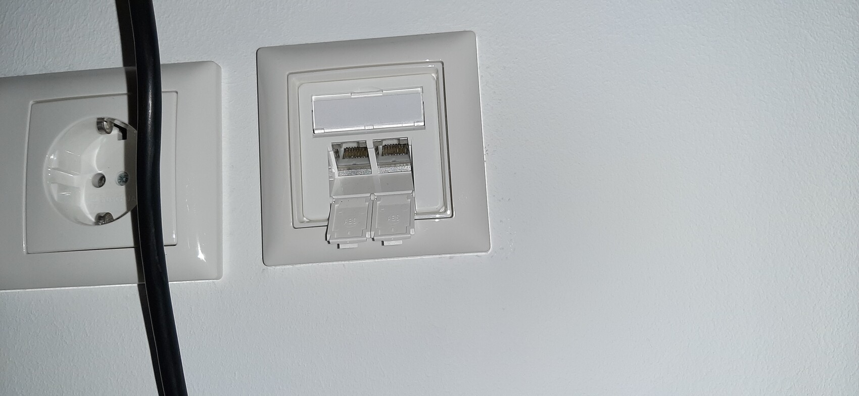Weiße Wandplatte mit zwei RJ-45-Netzwerkbuchsen und daneben runde Steckdose; schwarzes Kabel davor.