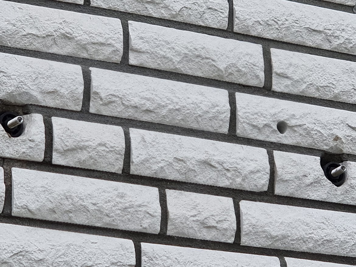 Rauer weißer Ziegelwand mit zwei schwarzen Öffnungen und herausstehenden Metallstiften.