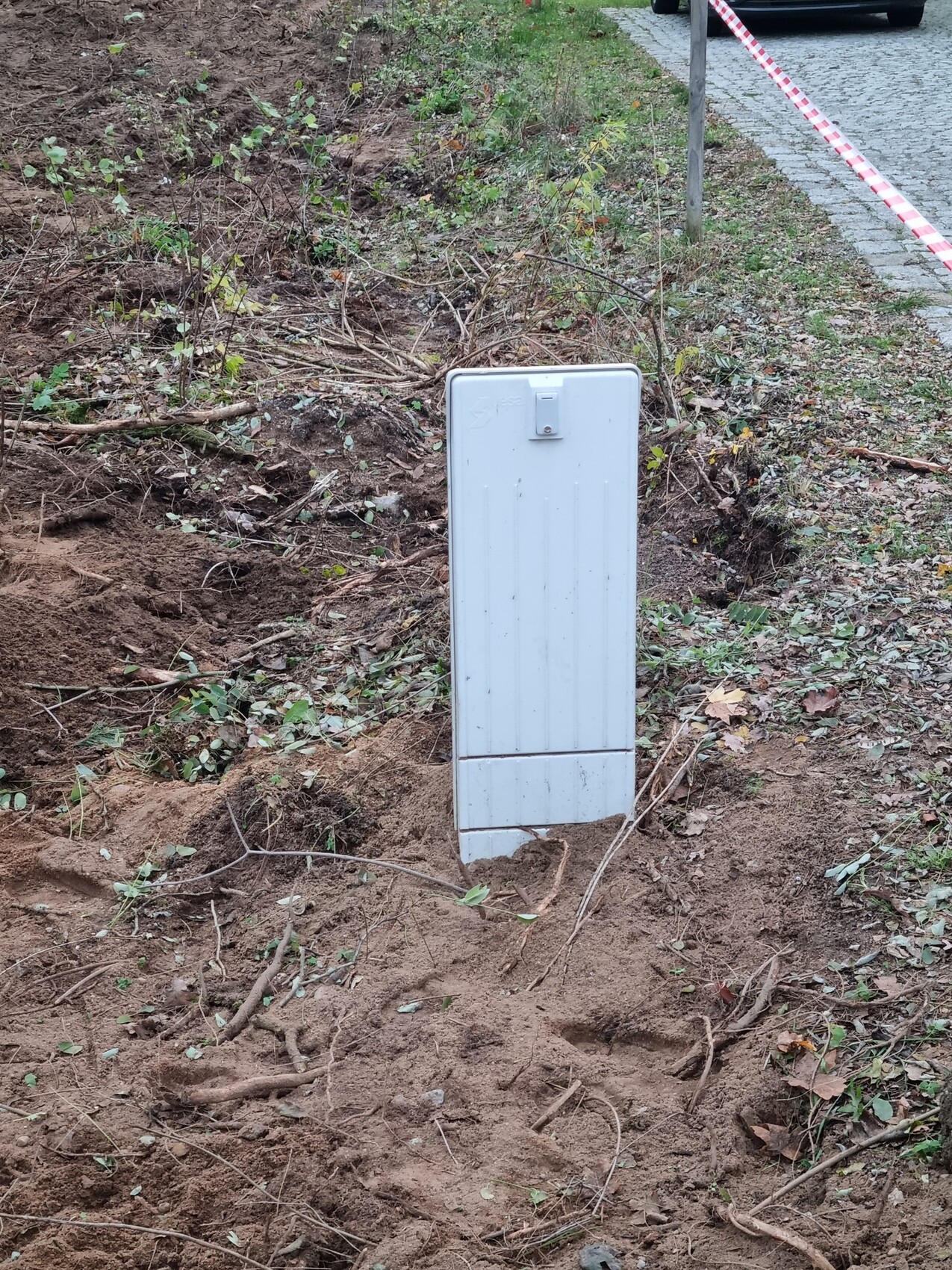 Weißer Metall-Verteilerkasten steht in offener Erdgrube, Absperrband im Hintergrund.
