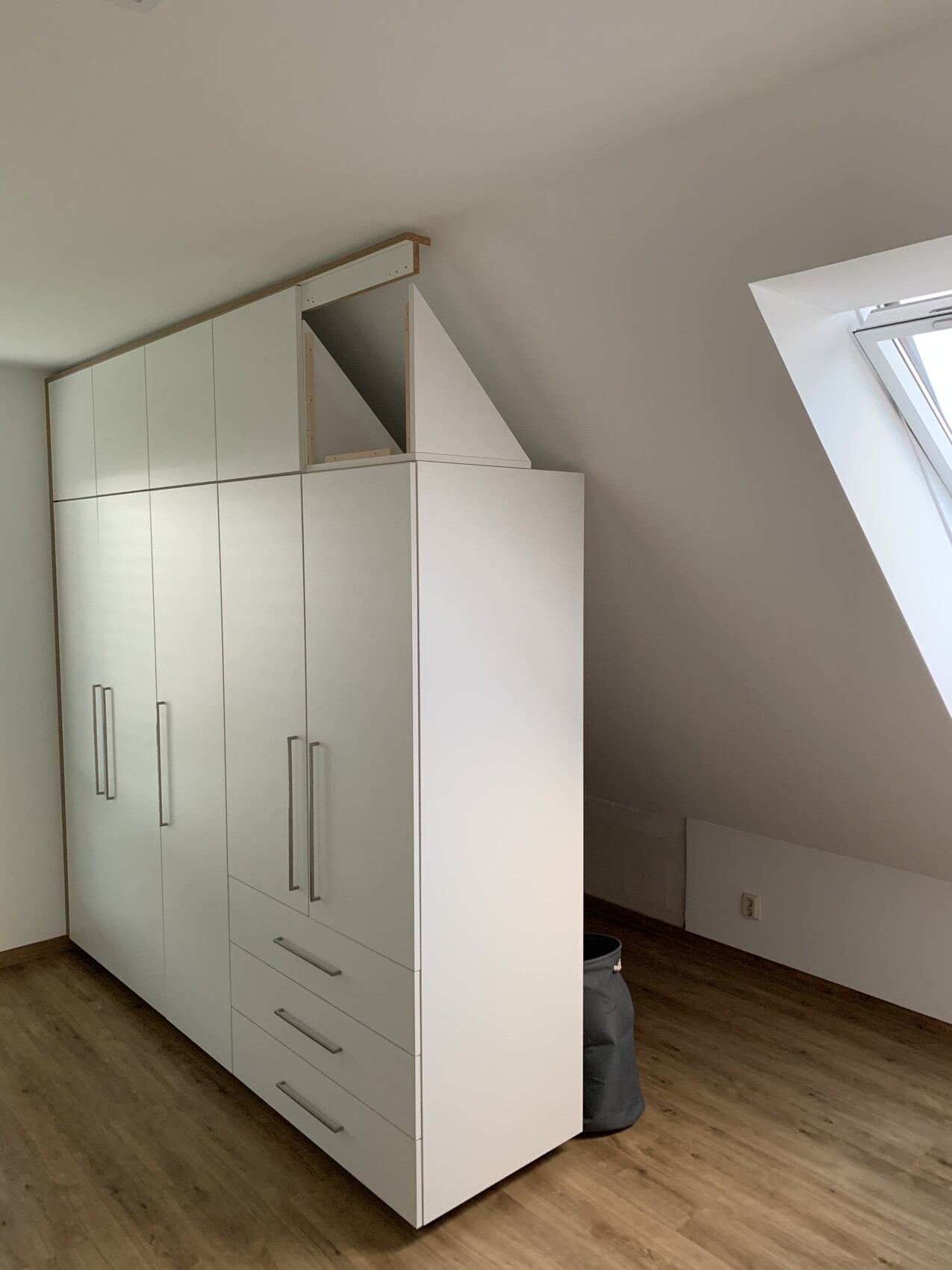 Weißer Kleiderschrank mit Türen und Schubladen unter Dachschräge im Dachgeschoss nahe Dachfenster.