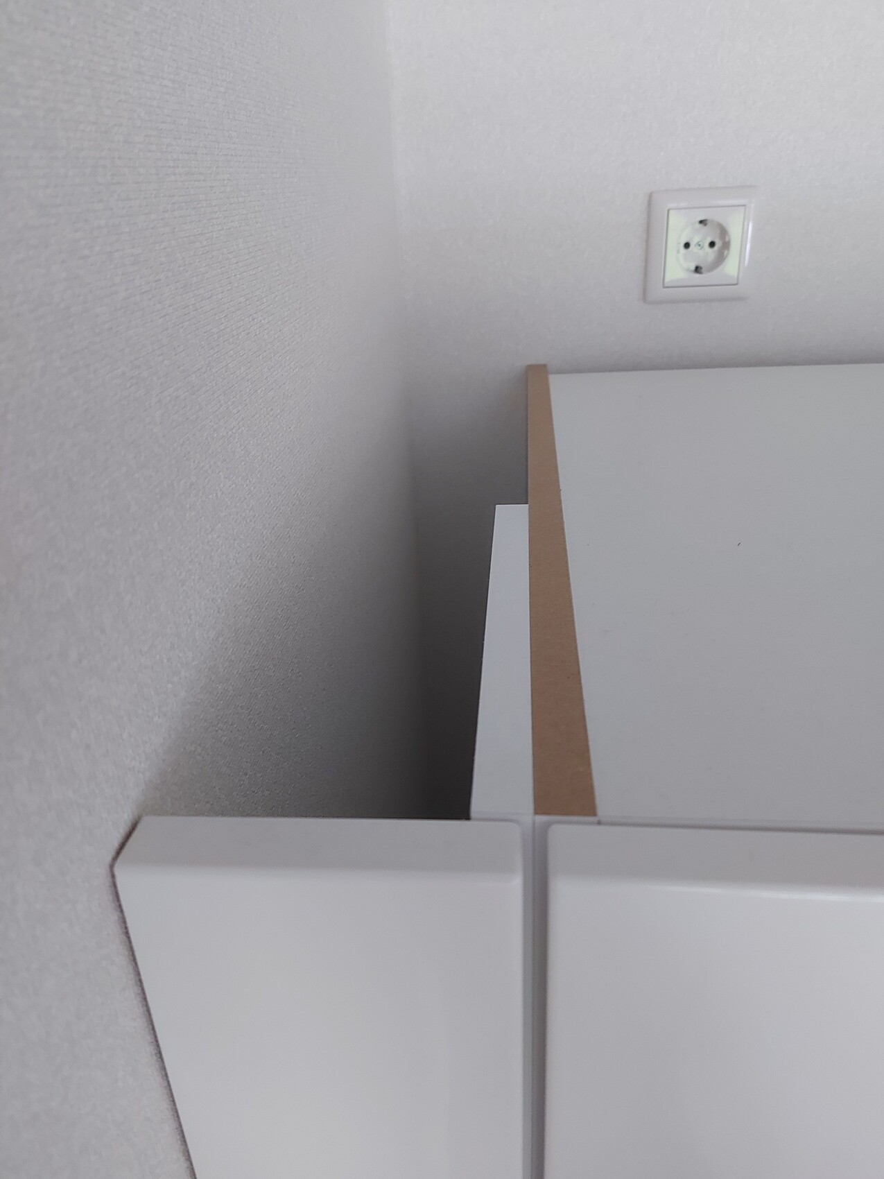 Eckansicht eines weißen Schranks mit brauner Kante vor grauer Wand; Steckdose sichtbar.
