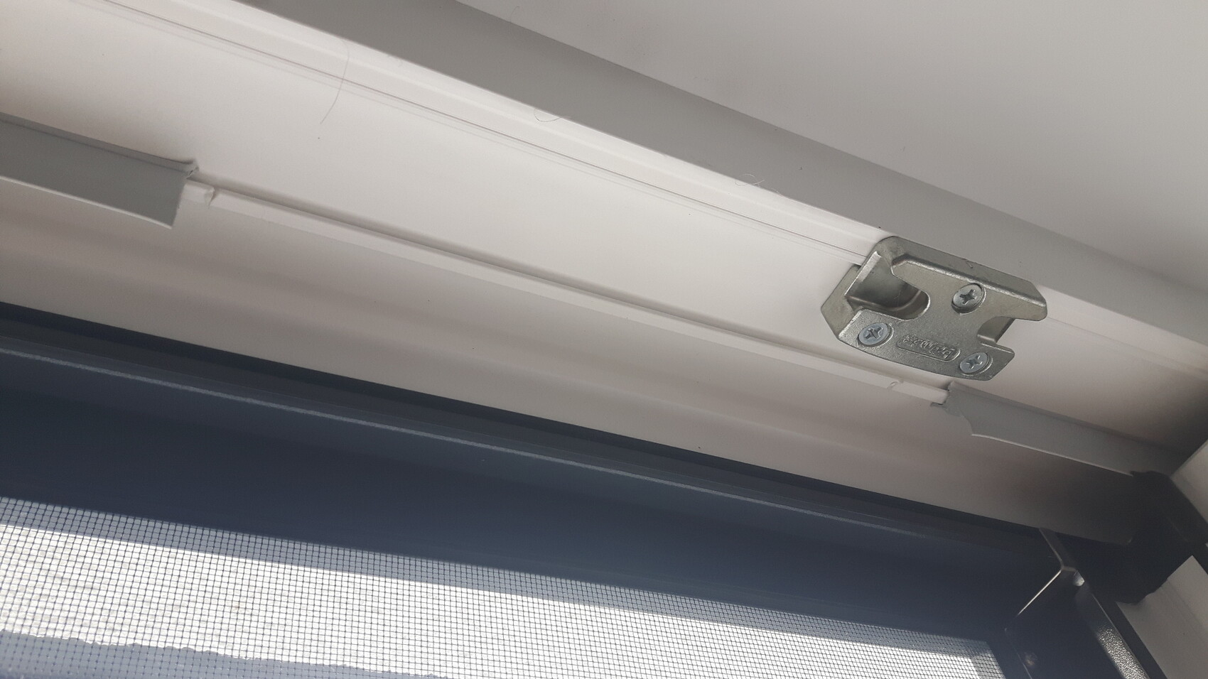 Metallische Halterung am weißen Fensterrahmen mit Fliegengitter darunter.