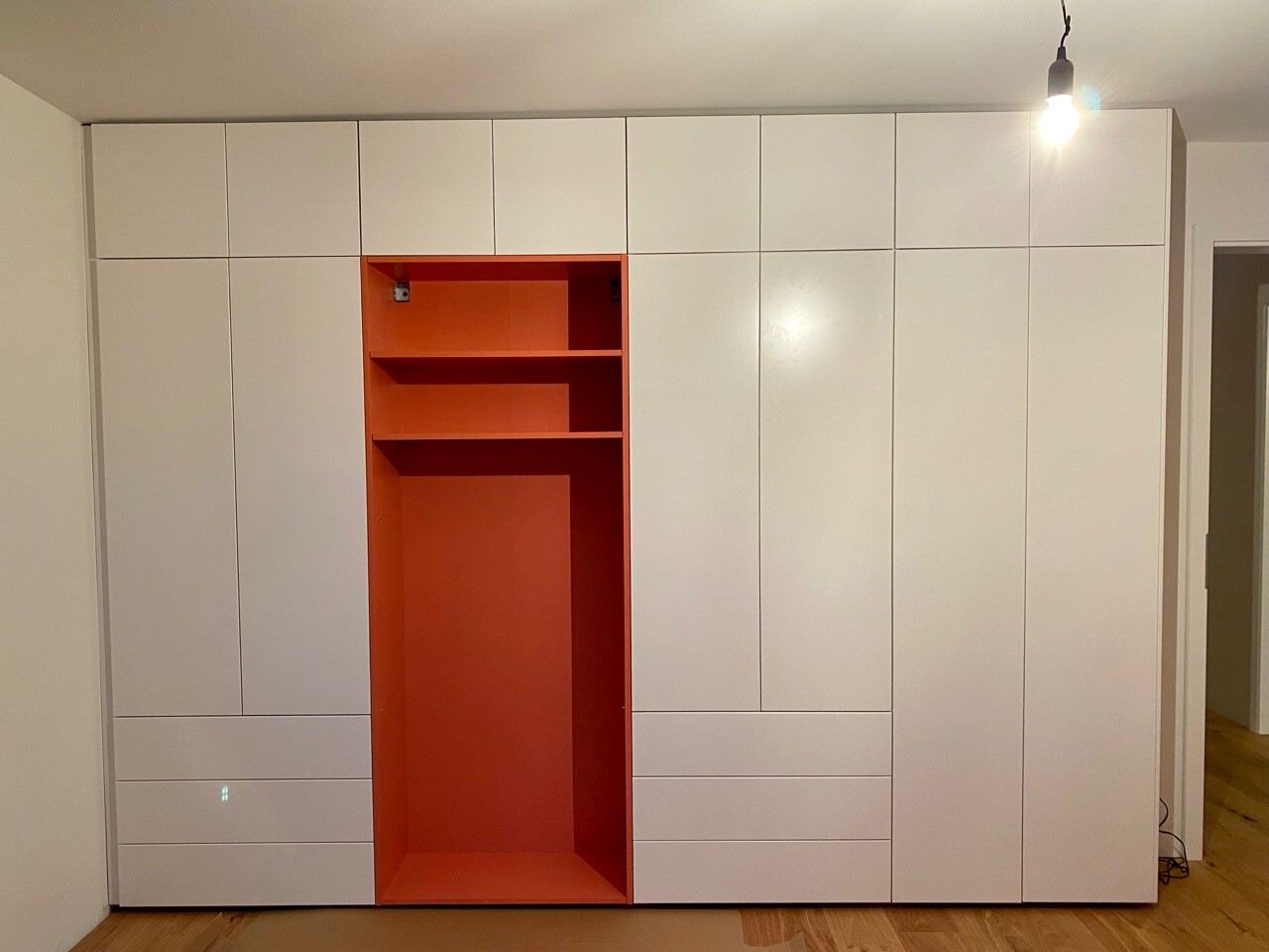 Weiße Einbauschrankwand mit orangefarbenem Innenraum in der Mitte, Regale und Schubladen, Holzboden