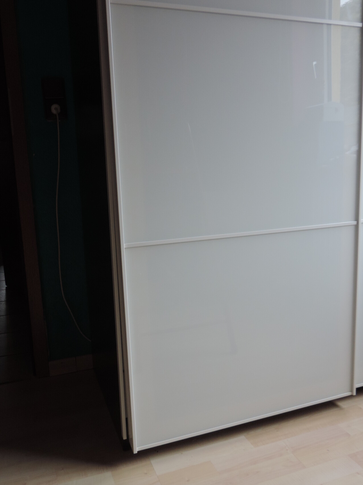 Großer weißer Schrank mit mattweißen Schiebetüren; links Steckdose mit Kabel, Holzboden sichtbar.