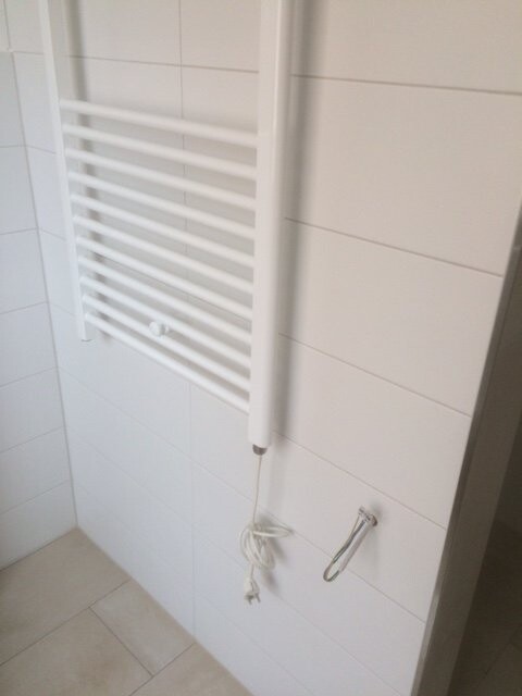 Weiße Handtuchheizung an einer Badezimmerwand, Kabel hängt herunter, Fliesen sichtbar