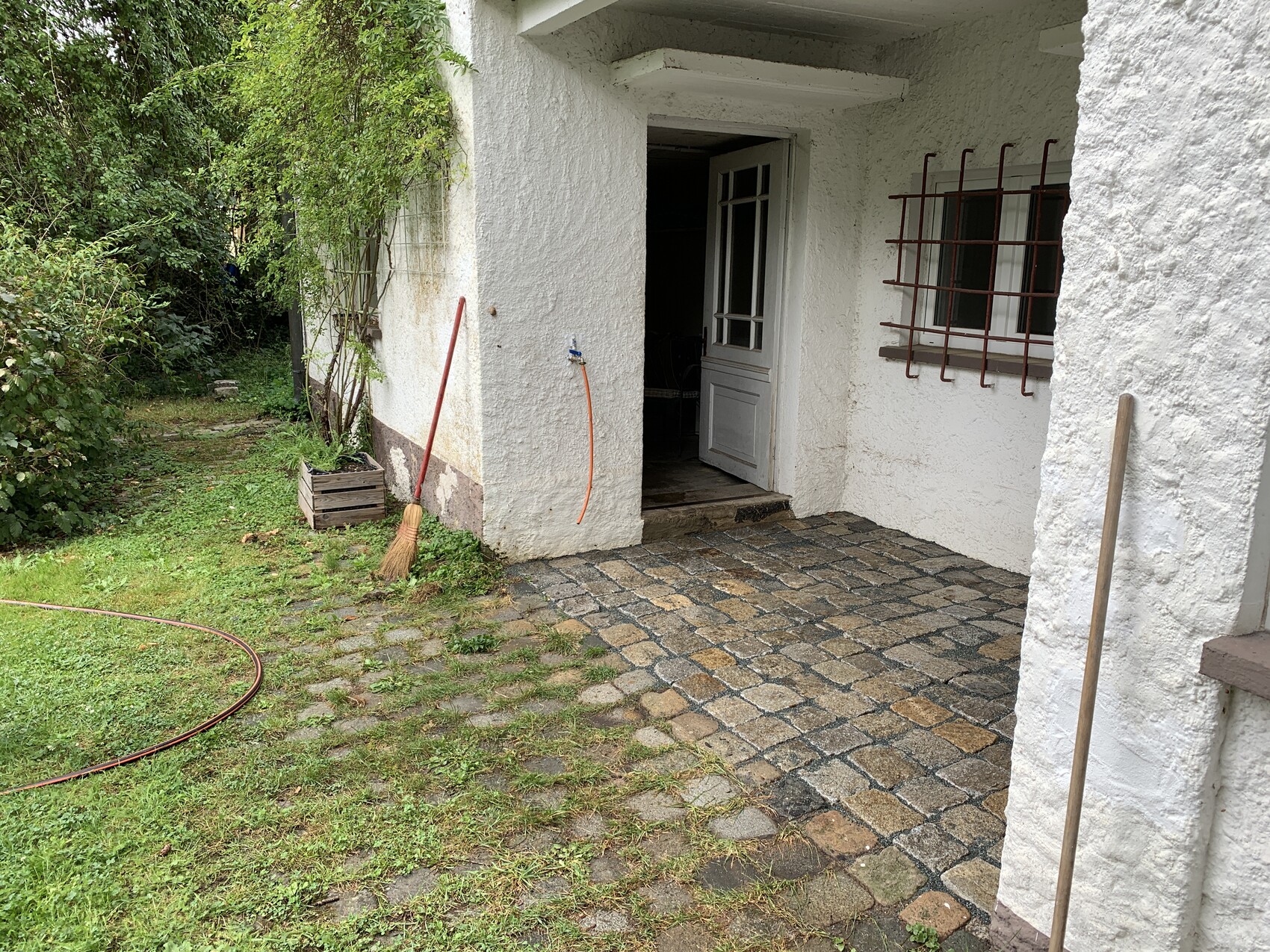 Weiße Hausfassade mit offenem Türportal, Fenster mit Rostgitter, Besen und Gartenschlauch.
