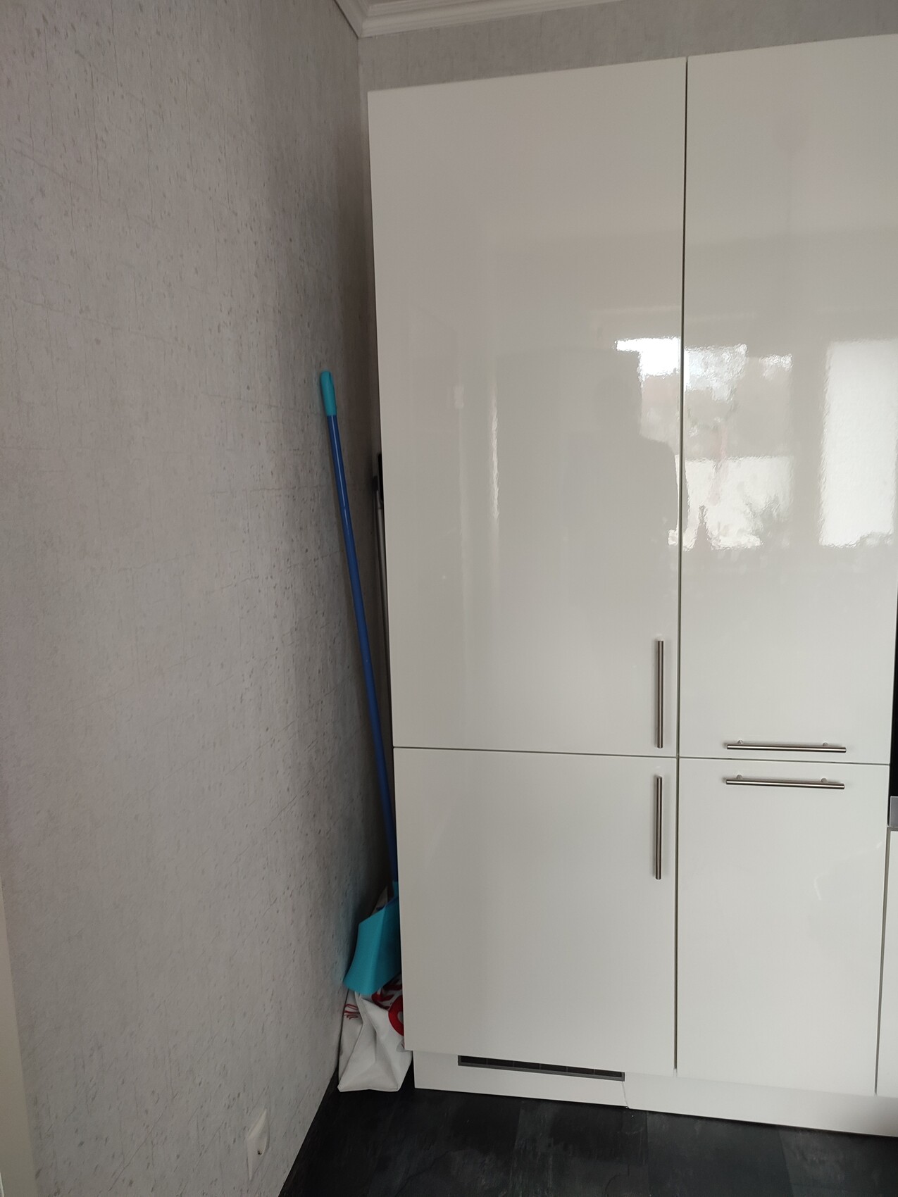Weiße Schränke in Abstellraum; blauer Reinigungsstiel lehnt an grauer Wand, Mülltüten am Boden.