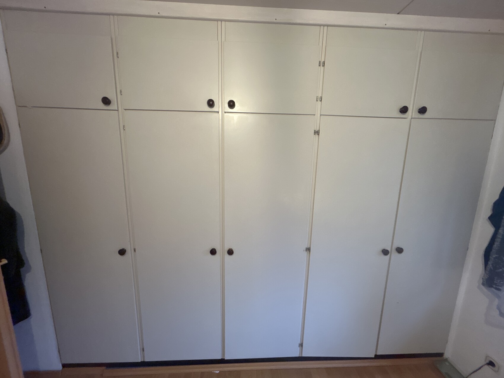 Weiße Schrankwand mit mehreren vertikalen Türen und runden dunklen Griffen; Holzfußboden sichtbar.