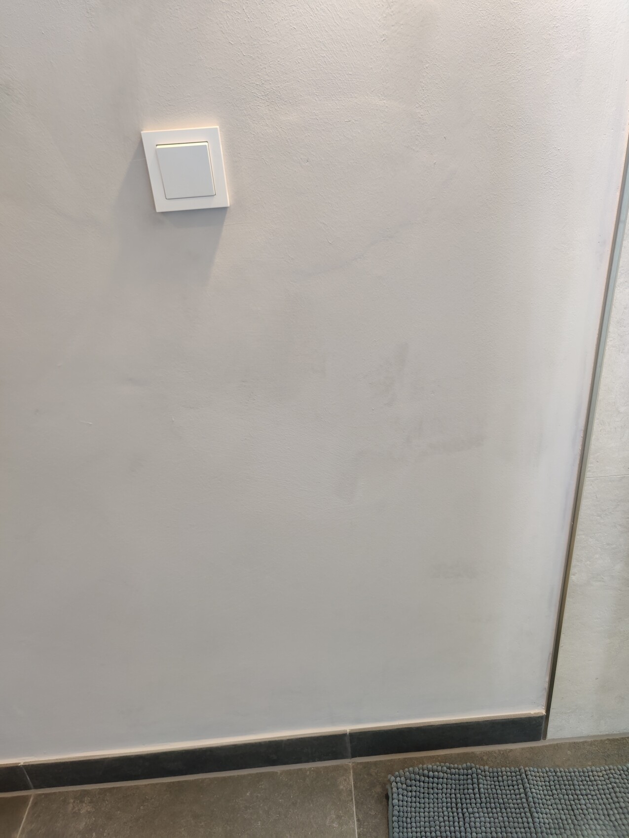 Weiße Wand mit quadratischem Lichtschalter; Schatten darunter, grauer Boden mit Matte.