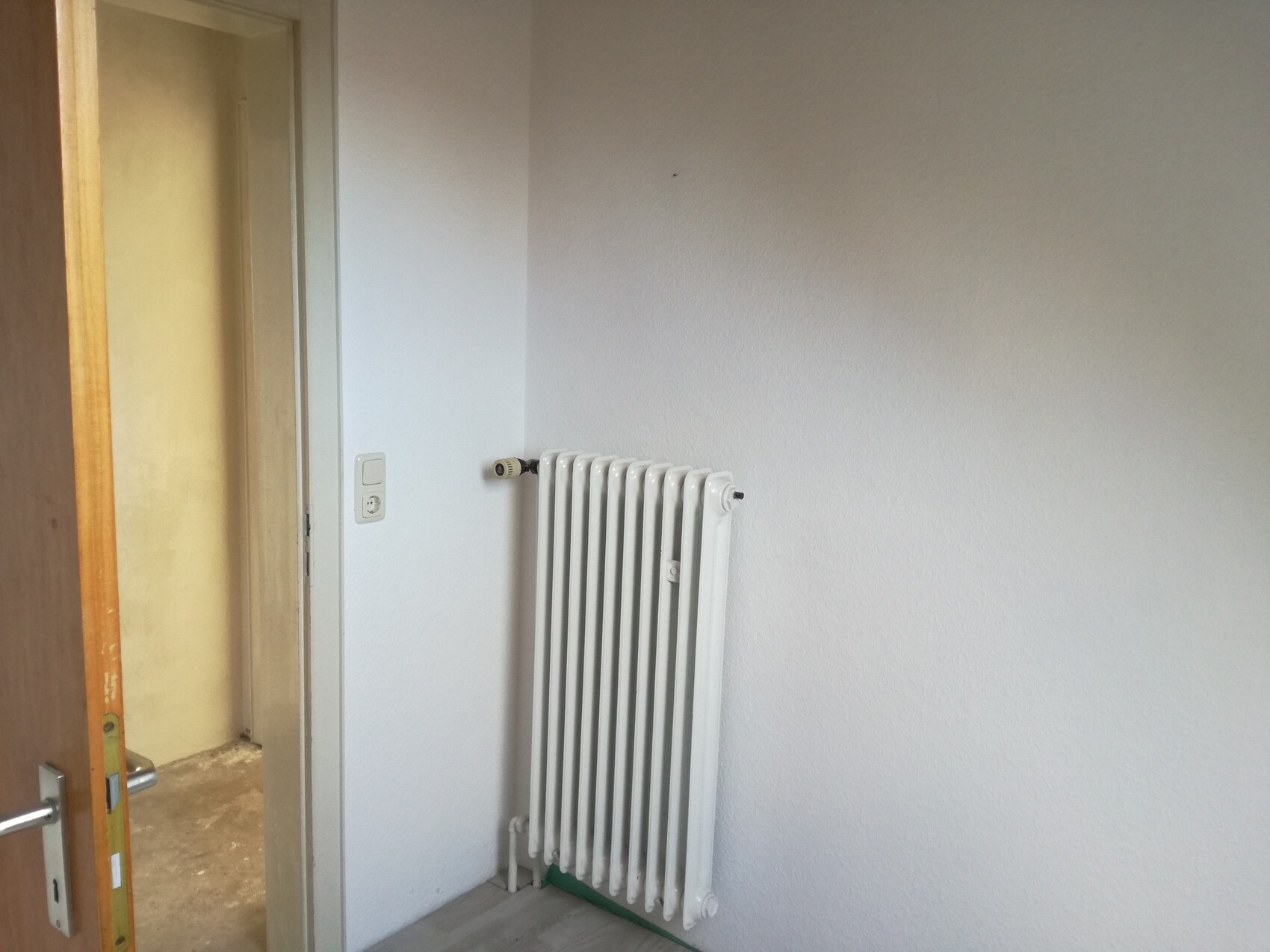 Weißer Rippenheizkörper an weißer Wand, Türrahmen links im Bild.