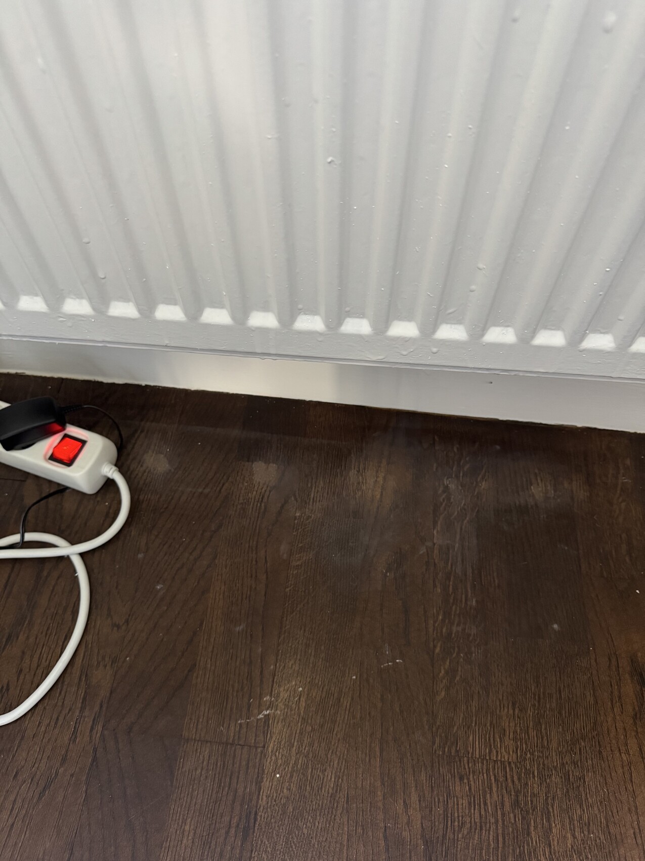 Weißer Heizkörper an der Wand, Steckdosenleiste mit rotem Licht auf dunklem Holzboden.