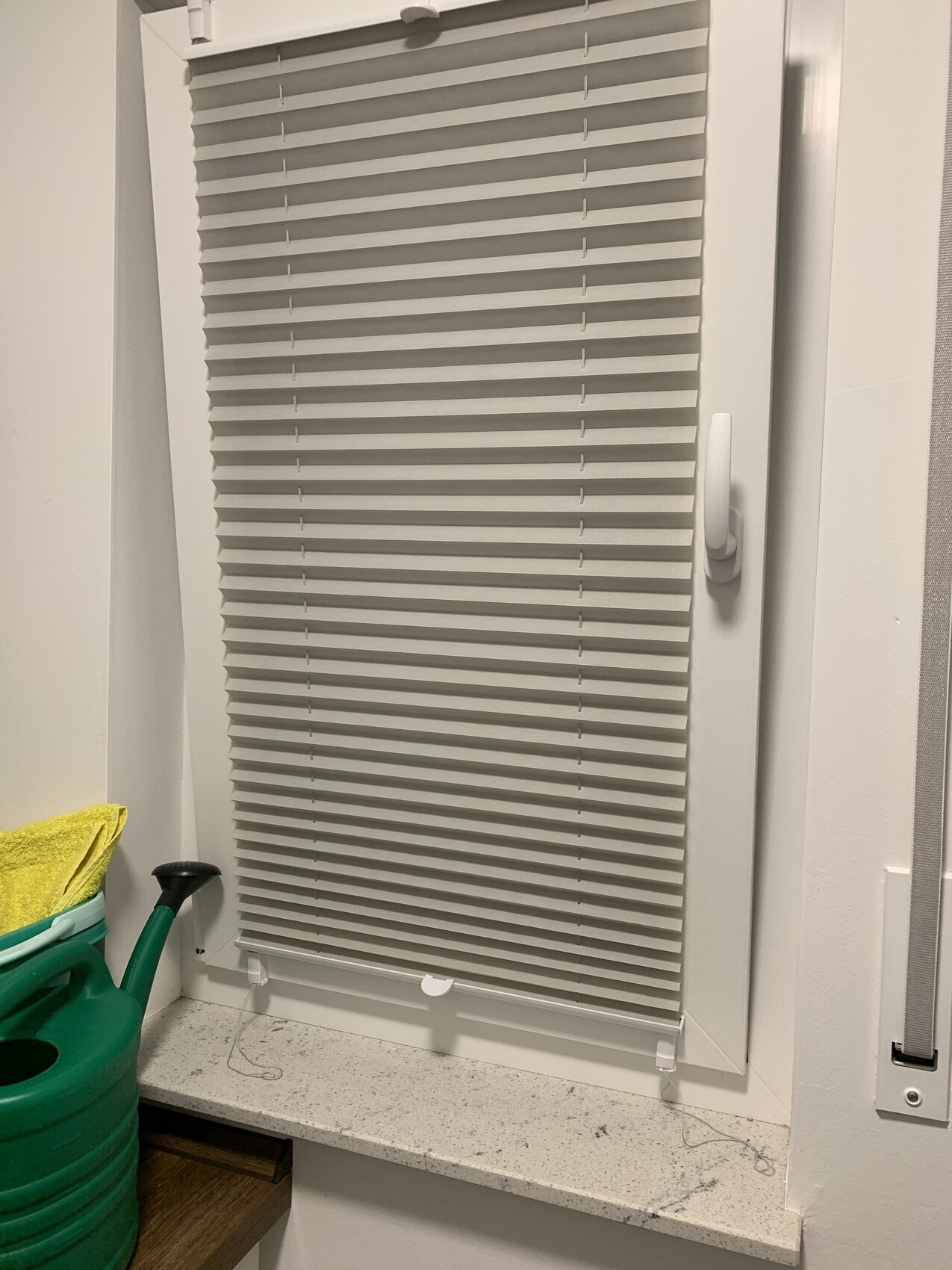Weiße Fensterjalousie mit geschlossenen Lamellen; grüne Gießkanne mit gelbem Tuch neben Fensterbank.