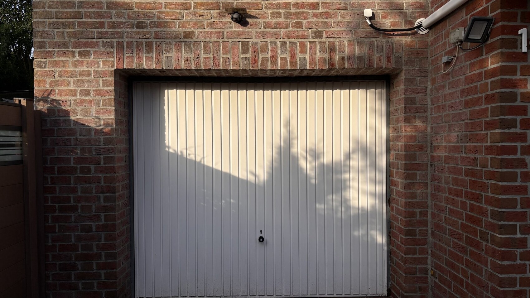 Weiße, vertikale Garagentür vor Backsteinwand; Baum-Schatten darauf, Außenkamera oben.