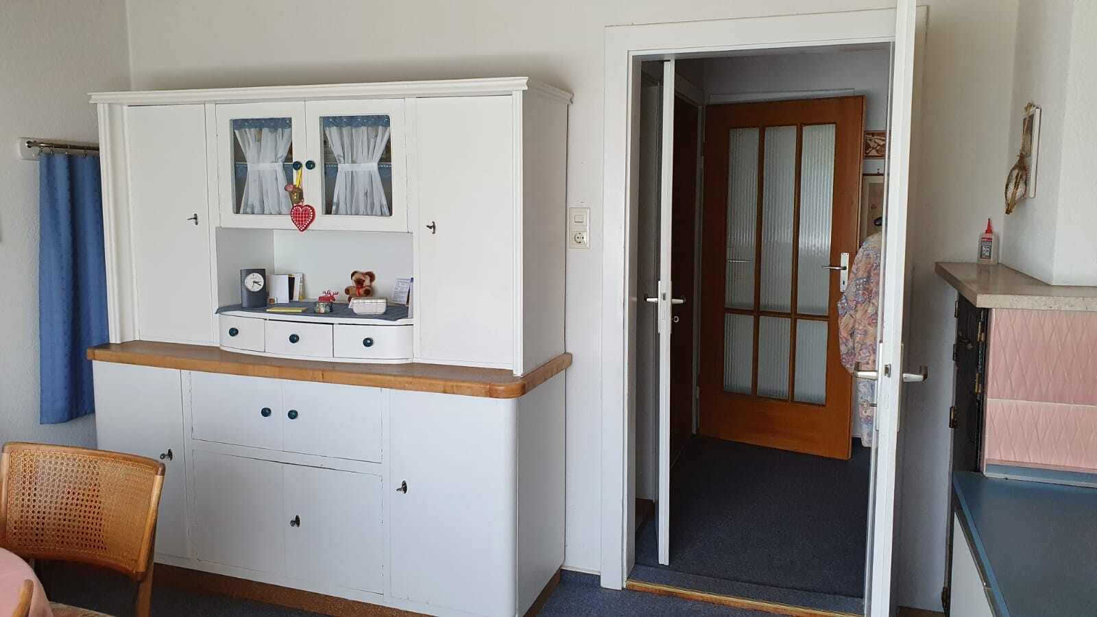 Helles Wohnzimmer: weißer Küchenschrank mit Glastüren, blauer Vorhang links, Tür zum Flur rechts.