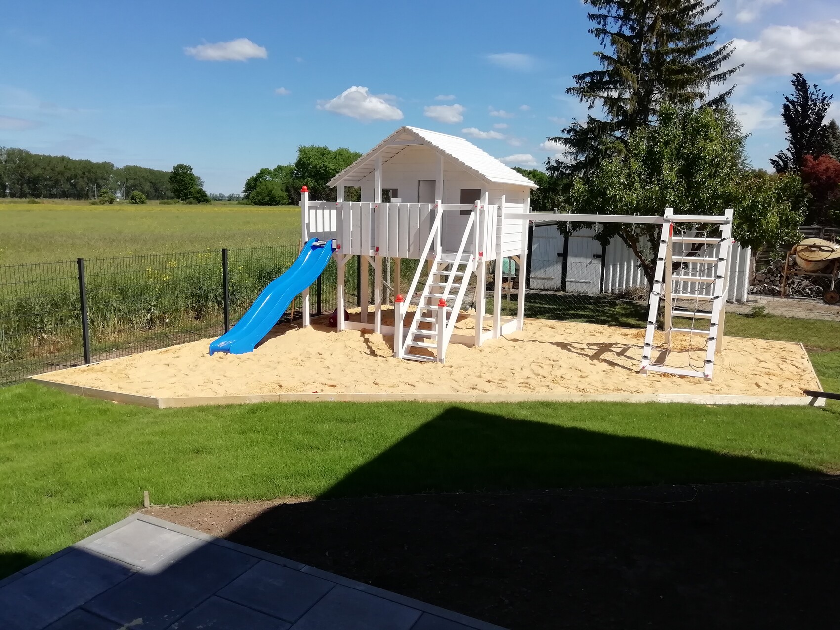 Weißes Spielhaus auf Sandkasten mit blauer Rutsche, Treppenstufen und Klettergerüst im Garten.