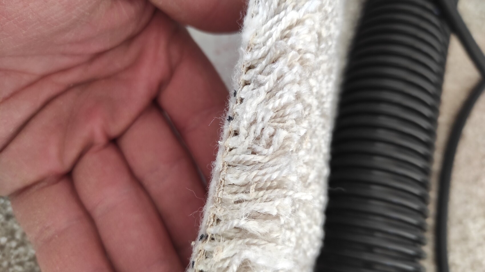 Nahaufnahme eines weißen Teppichrands mit Fasern, Hand links, schwarzer Spiralkabelkanal rechts.