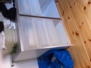 Weißer Unterschrank mit offenen Türen und Innenfächern, Holzfußboden, blaue Tüte daneben.