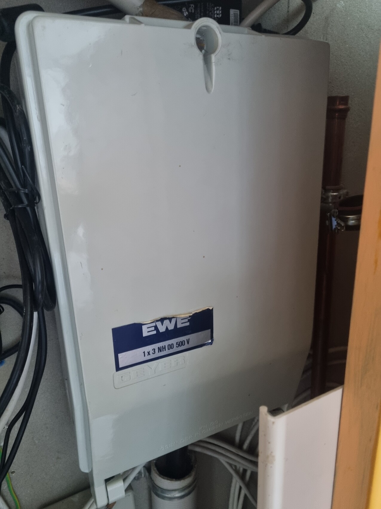 Weißes Elektro-Gehäuse mit blauem EWE-Aufkleber, Kabel links, Rohre rechts im Abstellraum.
