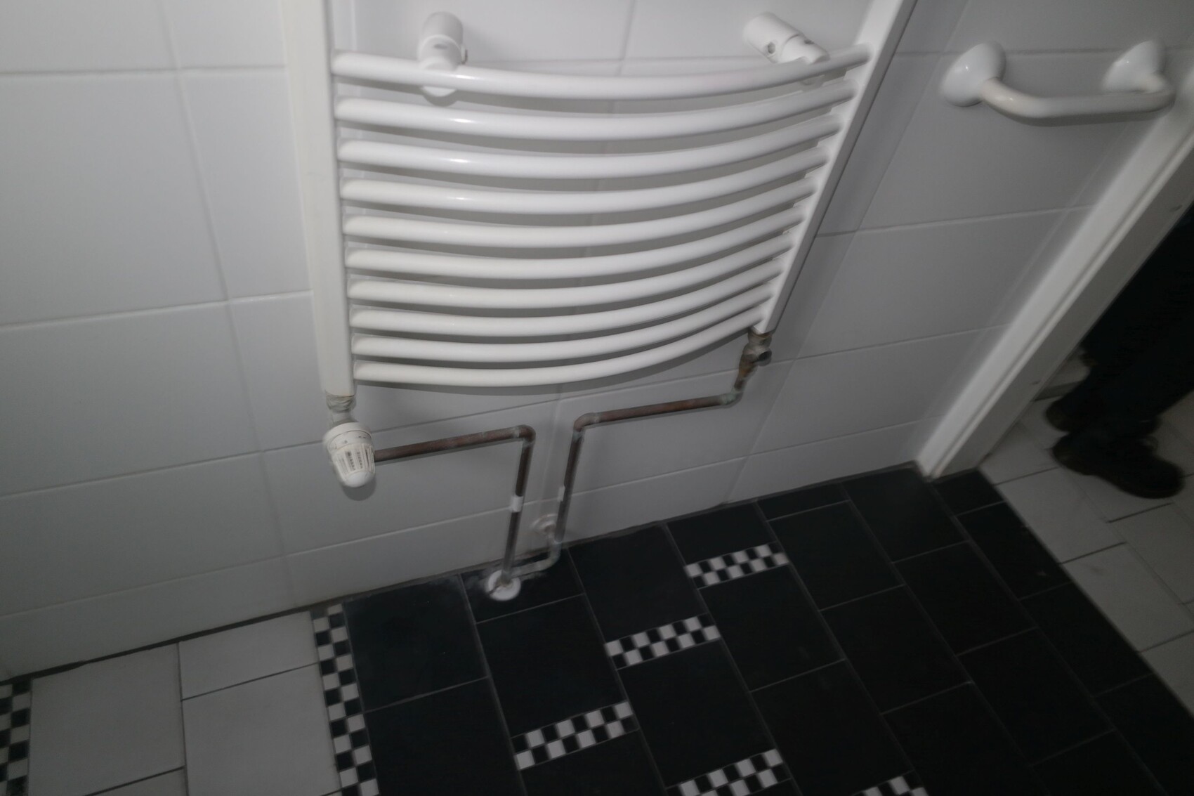 Weißer Handtuchheizkörper an Fliesenwand, Rohre nach unten, Boden mit Schwarz-Weiß-Fliesen.