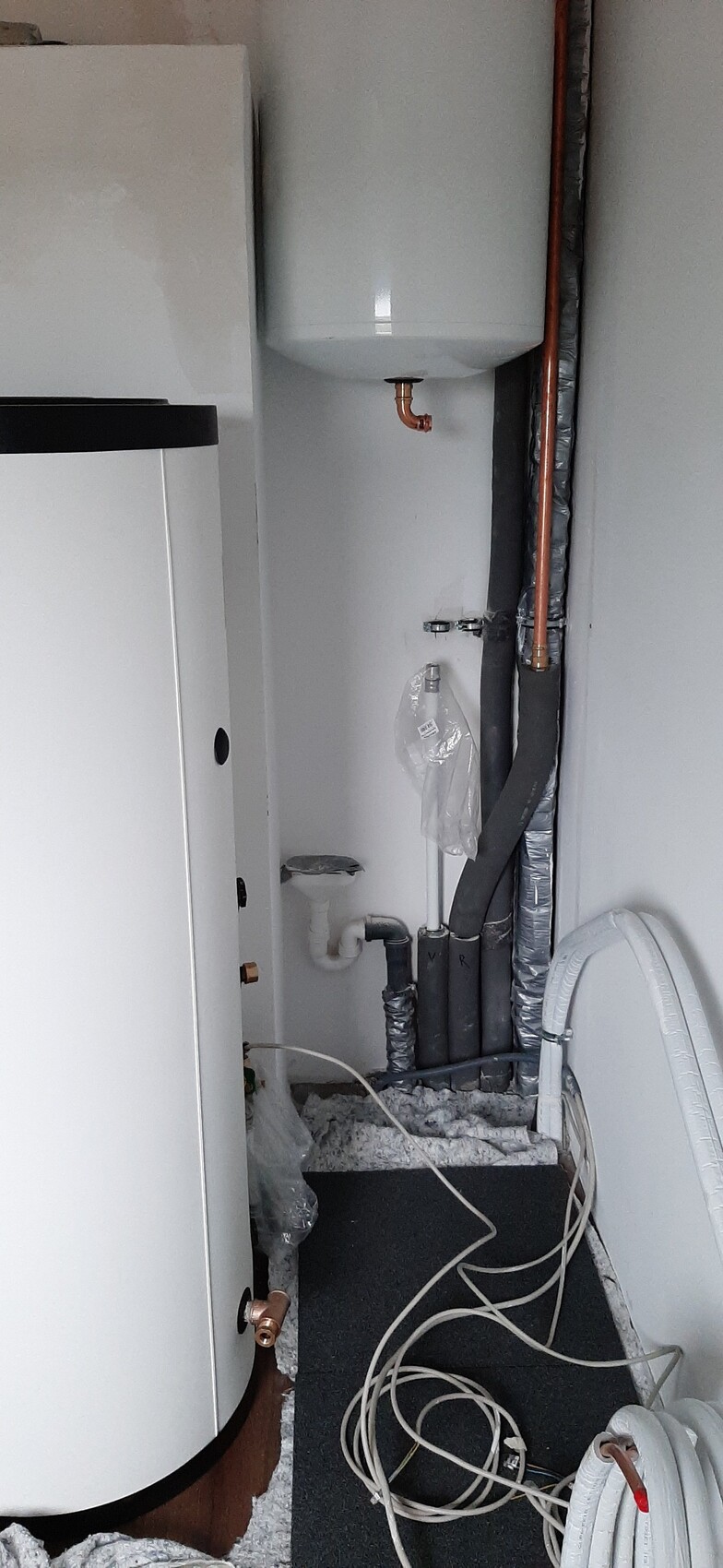 Heizungsraum mit weißem Warmwasserboiler links, Ausdehnungstank oben, Rohren und Kabeln am Boden.