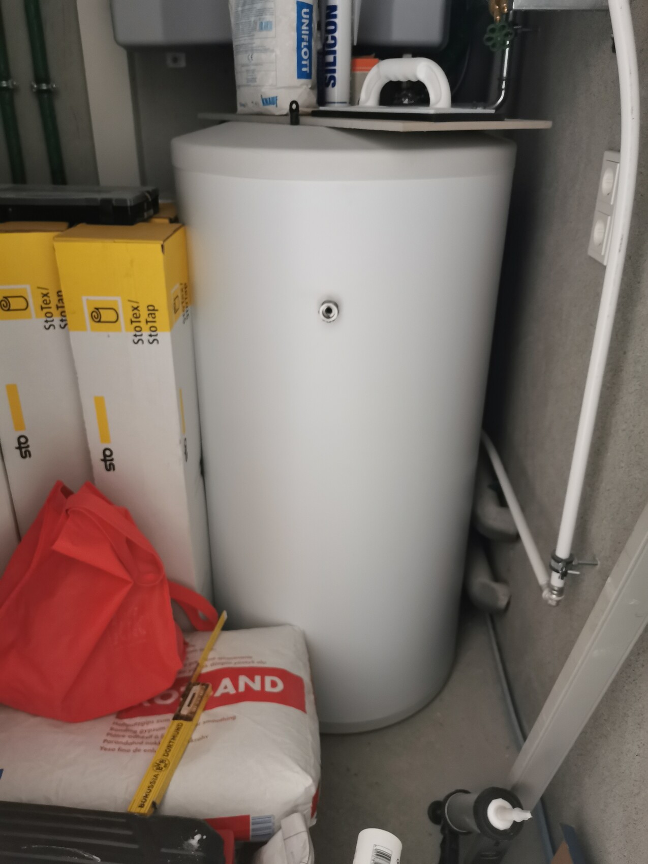 Weißer Warmwasserboiler in einem Abstellraum, umgeben von gelben Kartons und rotem Beutel.