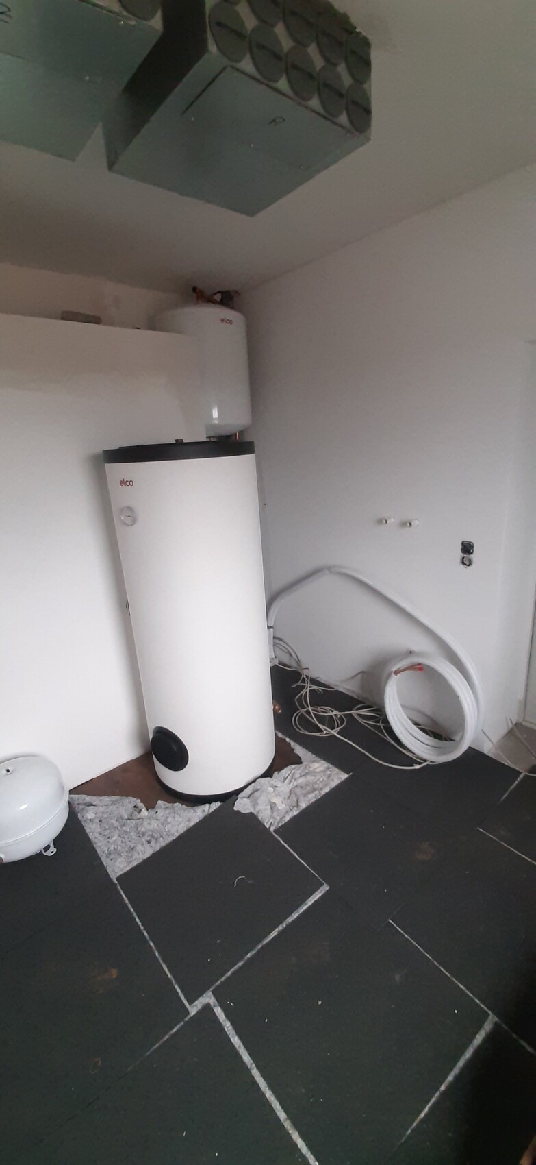 Weiße Wasserheizung/Boiler im Keller mit Ausdehnungstank, Kabeln und dunklen Fliesen.