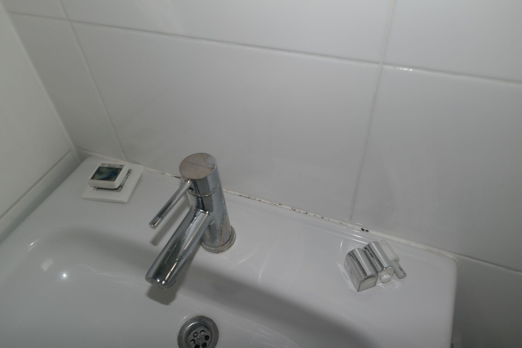 Waschbecken mit Chromarmatur; links kleine digitale Uhr auf Ablage, weiße Fliesen im Hintergrund.