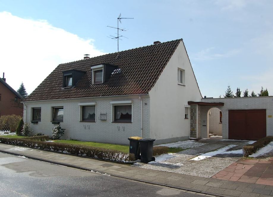 Weißes Einfamilienhaus mit Satteldach, Dachgaube, Garage, breiter Einfahrt und Mülltonnen davor.
