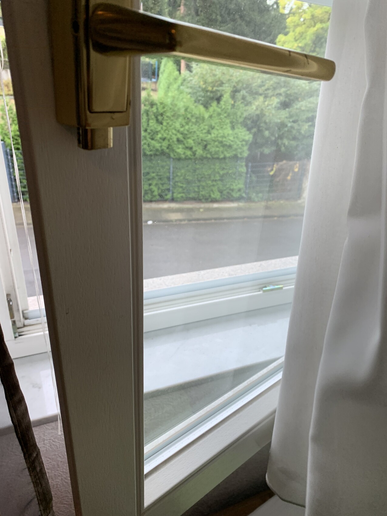 Fenster mit beigeem Griff, weiße Vorhänge, Blick auf nasse Straße und grüne Hecke.