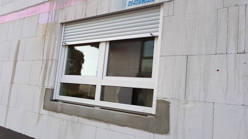 Fenster mit weißem Rahmen und Rollladen über grauer Betonwand; Putzreste sichtbar.