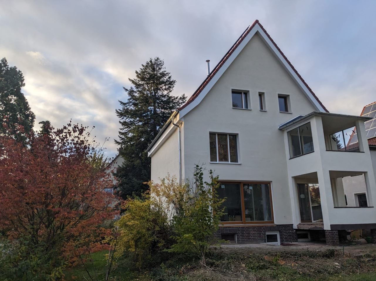 Helles zweigeschossiges Haus mit Balkon, großen Fenstern und Herbstbäumen davor.