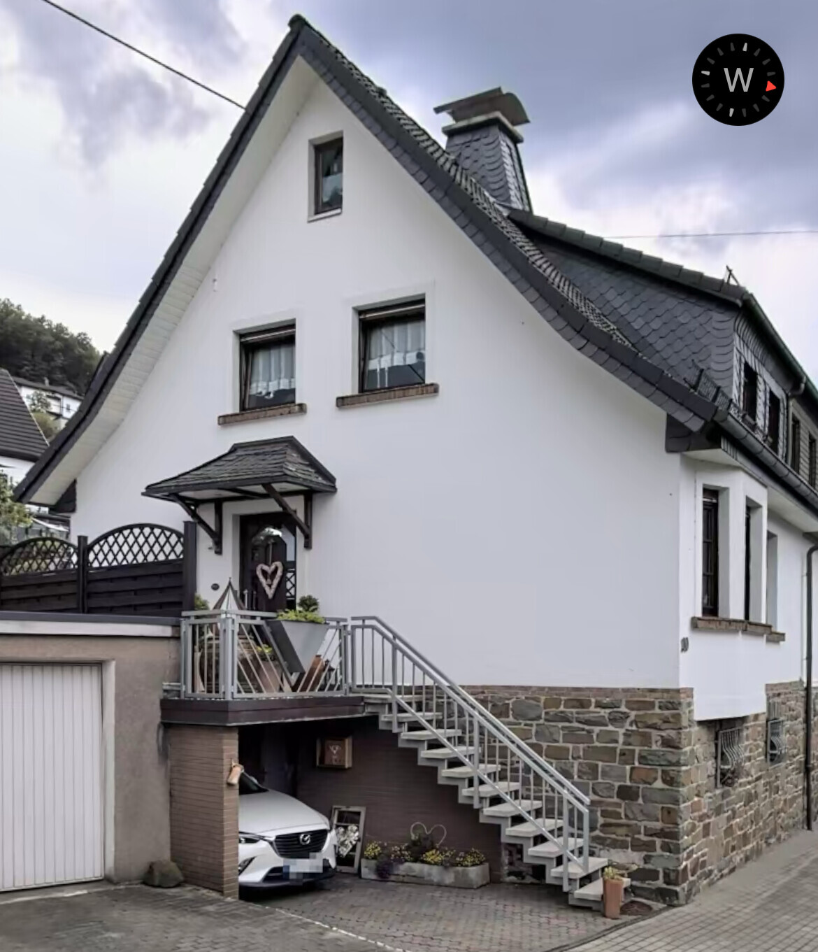 Weiße Fassade eines Hauses mit dunklem Schieferdach, Treppe zum Eingang, Auto darunter.